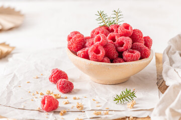 A rustic ceramic bowl filled with plump, fresh raspberries sits on a white table surrounded by...