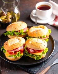 Four small ham and cheese sandwiches on buns, arranged on a dark plate.  A cup of tea and a small dish of olives are in the background
