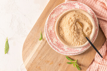 A close-up, top-down view of a bowl of a brown sugar a decorative plate with a spoon, a wooden board, fresh stevia leaves, and a pink napkin