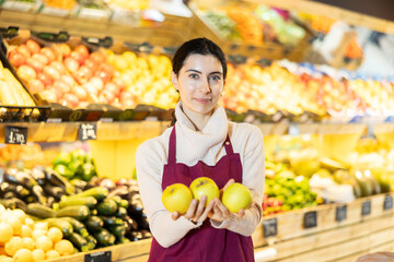 Woman staff with apple in hands, vendor demonstrate, adds and displays goods. In sales area of vegetable store, seller restructures display case.