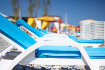 Horizontal travel defocused photo. Selective focus. Row of blue chaise longue, sun lounger, deck chair, colorful umbrellas, parasols, on pebble beach of Batumi. Black sea coast. Tourism, relax, calm