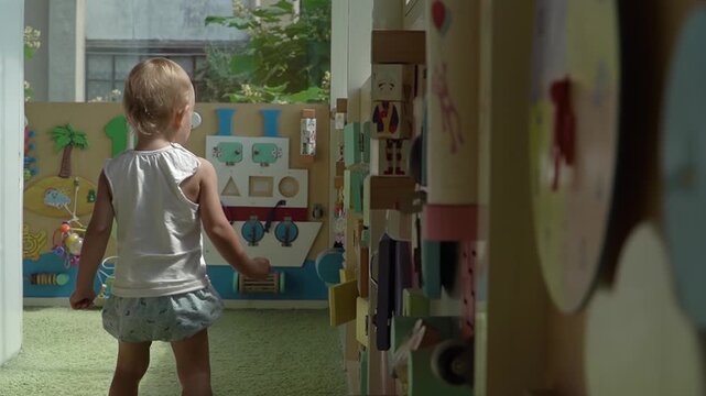 A baby girl with short blond hair and a beautiful blue suit eagerly and impatiently opens, closes, and twists colorful, different pieces of detail on a busy board all over the wall of the playroom.