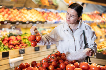 Positive middle-aged woman choosing tomatoes standing at counter in large vegetable market