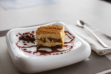 Delicious tiramisu dessert with cherries served on a white plate with a berry sauce. A square of tiramisu dusted with cocoa powder sits on a white plate, accompanied by a spoon on a table in cafe.