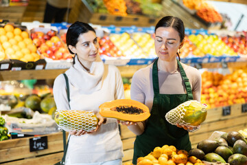 Armenian woman consults with a seller about choosing ripe papaya in a supermarket. Visitor to a vegetable store carefully chooses exotic fruits