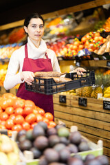 Female seller holds a plastic container with sweet potatoes in her hands against the background of racks with vegetables and fruits in a supermarket