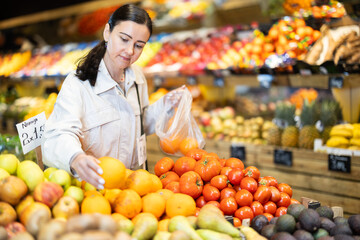 Focused positive middle aged woman shopping in organic food store, choosing ripe oranges