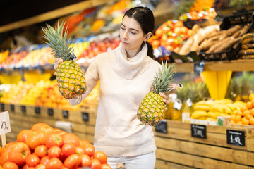 Woman chooses ripe pineapples against the background of shelves with vegetables and fruits in a supermarket