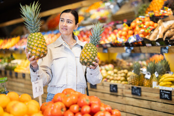 Positive middle-aged woman choosing pineapple standing at counter in large vegetable market