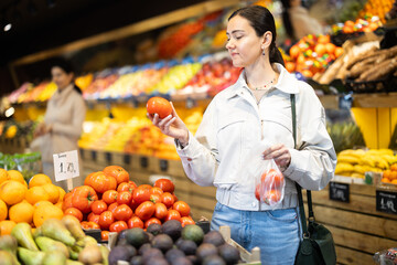 In vegetable store, young woman customer buy ripe tomato. Vegetables and fruits from different parts of world, products from local farms.