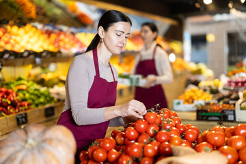 Vegetable shop assistant working in greengrocery. Woman seller in sales area with tomato. Employee demonstrates supply of fruit. Local and imported farm vegetables and fruits