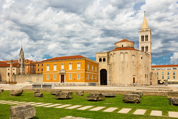 Fototapeta premium Stone Church of St. Donatus and tall bell tower rising next to remains of Roman forum in old Croatian city of Zadar, reflecting ancient historical and cultural roots under cloudy summer sky