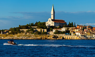 Naklejka premium Rare sailboats and motorboats float past coastal cliffs. Bell tower of Church of St. Euphemia rises above city of Rovinj, citys dominant feature.