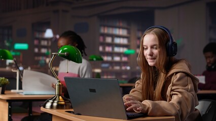 Girl studying in the library to obtain her diploma with focus music on headset, using the academic...