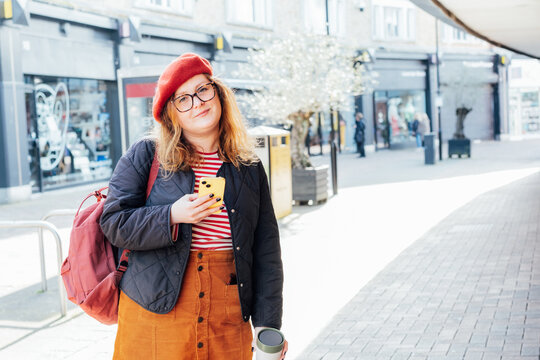 Young smiling plus size woman with backpack using mobile phone, drinking coffee outdoors on the street. Confident fashion hipster messaging, chatting, working remotely. Using navigator app for travel.