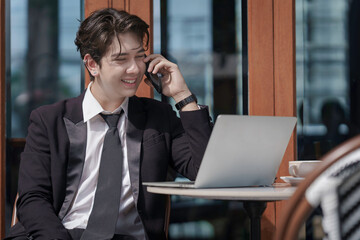 A young businessman in a black suit works at a coffee shop, using a laptop and a mobile phone while drinking coffee outdoors. The New technology business works.