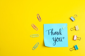 Blue sticky note with “Thank you” text surrounded by pastel binder clips and bright paper clips on a vivid yellow background, symbolizing gratitude and cheerful office creativity.