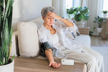 Worried senior woman checking blood pressure at home with digital monitor on sofa