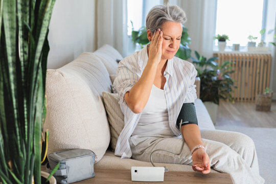 Senior woman with headache measuring blood pressure at home, sitting on cozy sofa