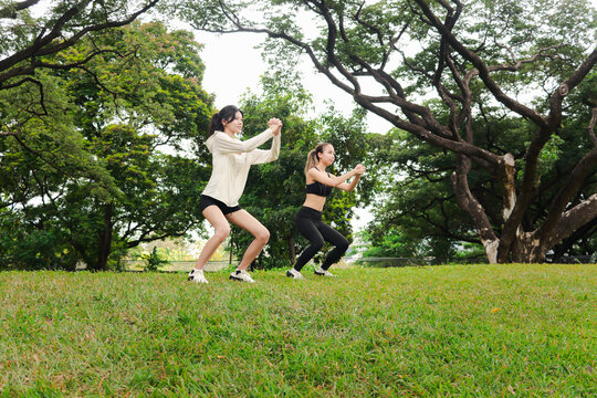 Two Asian women in sportswear stretching body outdoor in green park together. Sporty buddy women work out with squats in green garden. Sporty girl engage in healthy exercise with partner in nature