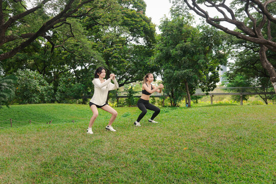 Two Asian women in sportswear stretching body outdoor in green park together. Sporty buddy women work out with squats in green garden. Sporty girl engage in healthy exercise with partner in nature