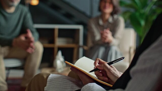 Hands of female psychologist taking notes in notebook while listening to couple during therapy session in clinic. Close-up view