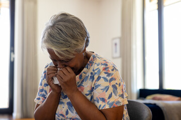 Senior African American woman sitting on chair in floral top dabbing nose with tissue by bed
