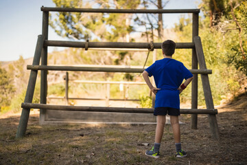 Fototapeta premium Boy standing with hands on hips facing large wooden log obstacle frame in sunlit forest clearing