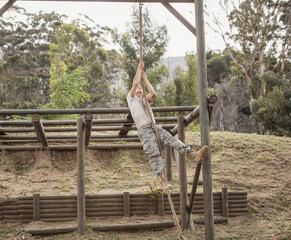 Male military trainee wearing camo climbing rope on wooden frame obstacle course in grassy clearing