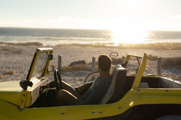 Man sitting in yellow dune buggy on sandy beach, gazing toward ocean horizon with bicycle