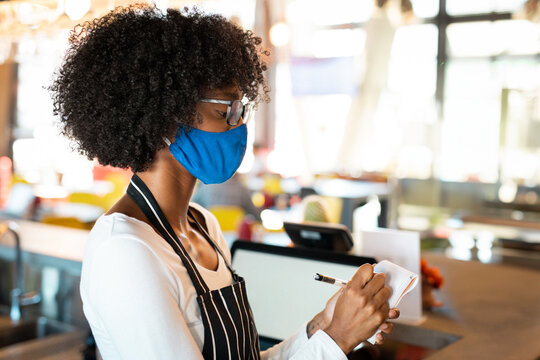 African American woman in apron, mask, taking order on notepad behind cafe register, copy space - Powered by Adobe
