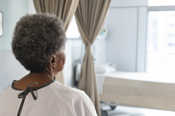 Senior African American patient wearing gown sitting in ward facing bed, viewing window, copy space