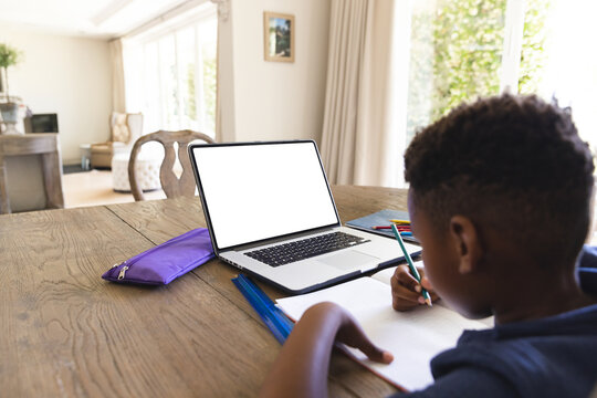 African American boy writing in notebook with pencil at table in room with laptop, copy space - Powered by Adobe