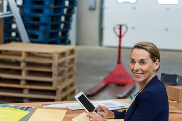 Warehouse supervisor sitting at desk holding tablet and reviewing charts near red pallet jack