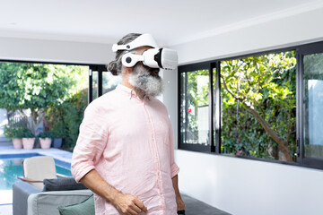 Senior man using virtual reality headset in bright living room with gray sofa, sliding glass doors