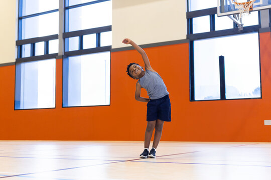 African American boy stretching in sportswear on gym court lines by windows and hoop, copy space