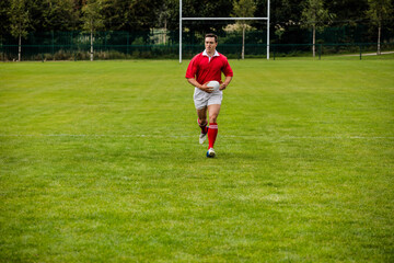 Male rugby player wearing red jersey sprinting while holding white ball on pitch before goalposts