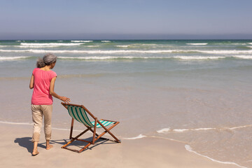 Naklejka premium Senior African American woman positioning striped beach chair on wet sand at ocean edge, copy space