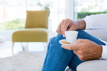 Hands are holding small white coffee cup with saucer and biscuit on sofa near large windows