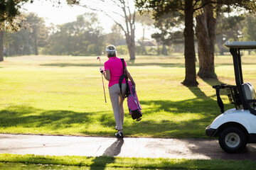 Mature female golfer walking along fairway path in pink polo shirt carrying bag and holding club