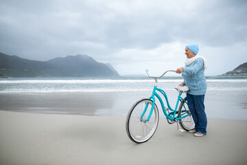 Naklejka premium Senior woman holding teal bicycle on beach wearing beanie scarf slip-ons gazing at sea, copy space