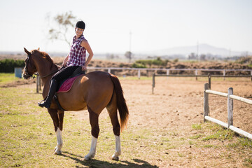 Female rider sitting astride chestnut horse in dirt paddock wearing helmet with burgundy saddle pad