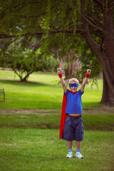 Boy standing on grassy park lawn, raising arms, wearing red cape, blue mask, red wristbands, bench
