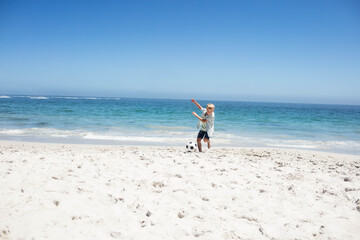 Naklejka premium Boy kicking soccer ball at shoreline on white sand near turquoise ocean under clear sky