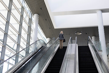 Woman riding escalator in modern lobby featuring glass wall, white columns, slatted ceiling