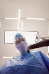 Obraz premium Female medical professional leaning beneath surgical light wearing mask, face shield in sterile OR