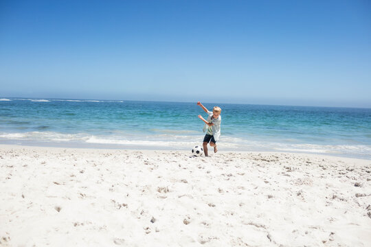 Boy kicking soccer ball at shoreline on white sand near turquoise ocean under clear sky