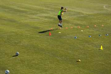 Male athlete wearing vest doing plyometric drill on field with cones and soccer ball, copy space