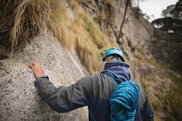 Senior male climber scaling mountain face carrying rope on backpack wearing teal helmet and jacket