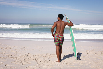African American man standing shirtless on beach holding surfboard facing rolling ocean waves